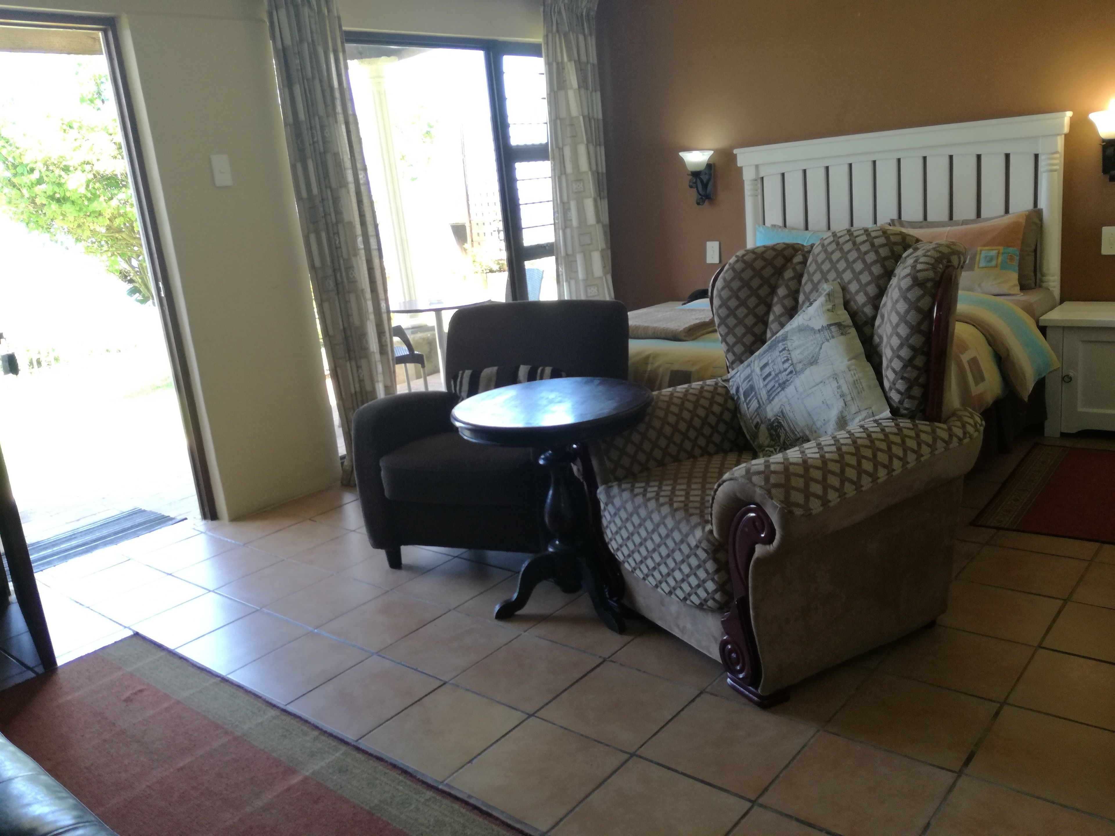 A room with a sofa, a small round table, a bed with patterned bedding, a nightstand, and a wall-mounted lamp.