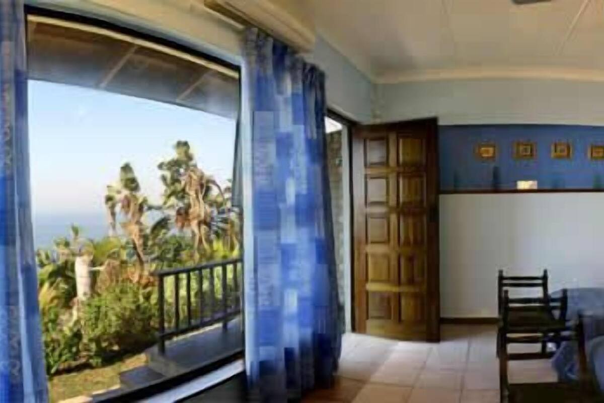 A bedroom with two beds, a wooden chair, a nightstand with a lamp, and a view of the outdoors.