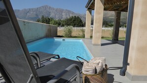 A pool area with lounge chairs and a towel basket, overlooking mountains. - Saddlebrook Cottage (Franschhoek)