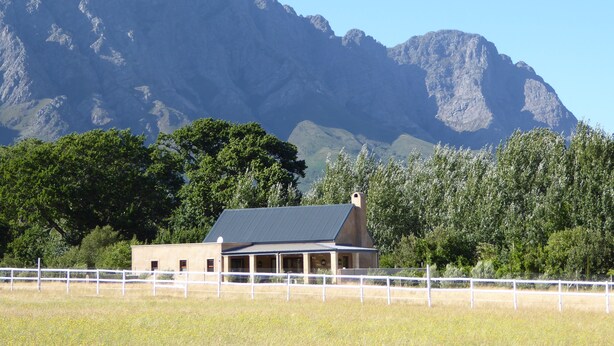 A building with a sloped roof, surrounded by a white fence, set against a backdrop of mountains and trees. - Saddlebrook Cottage (Franschhoek)