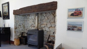 A stone fireplace with a modern wood-burning stove, a black piano, and a framed painting on the wall. - Saddlebrook Cottage (Franschhoek)