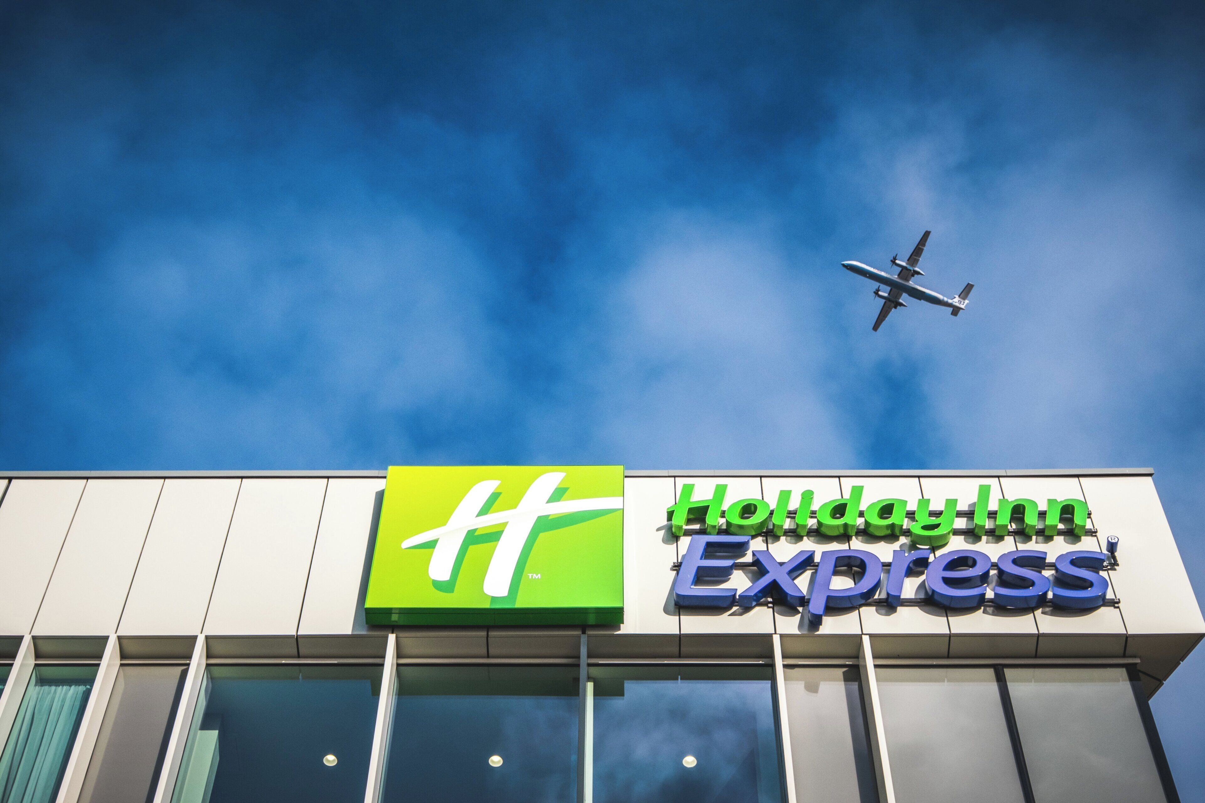 Ein Holiday Inn Express Schild vor einem wolkenlosen, blauen Himmel, im Hintergrund ist ein Flugzeug zu sehen.