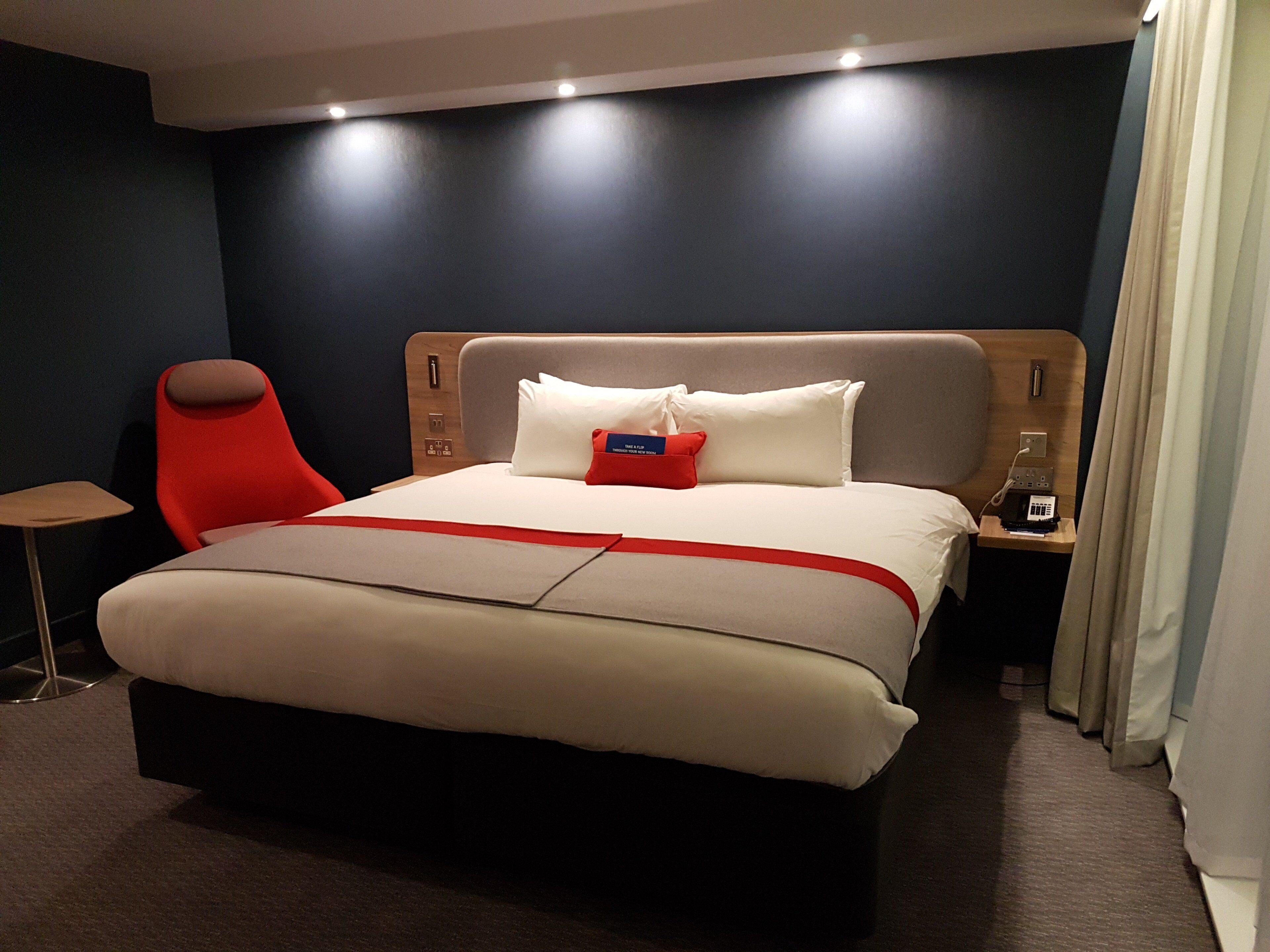 A modern hotel room with a large bed, a red chair, a bedside table with a phone, and a nightstand with a lamp.