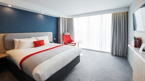 Une chambre d’hôtel moderne avec un grand lit, un fauteuil rouge, une petite table et une grande fenêtre avec des rideaux.