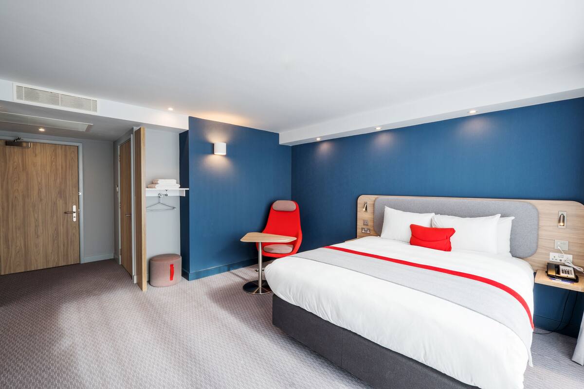 A modern hotel room with a large bed, a red chair, a small table, and a wooden nightstand.