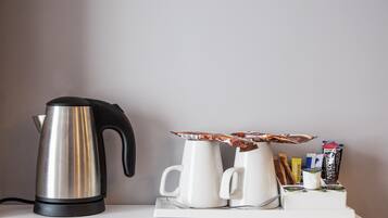A stainless steel kettle, two white mugs, and a coffee pod holder with various coffee pods on a white table against a plain wall.