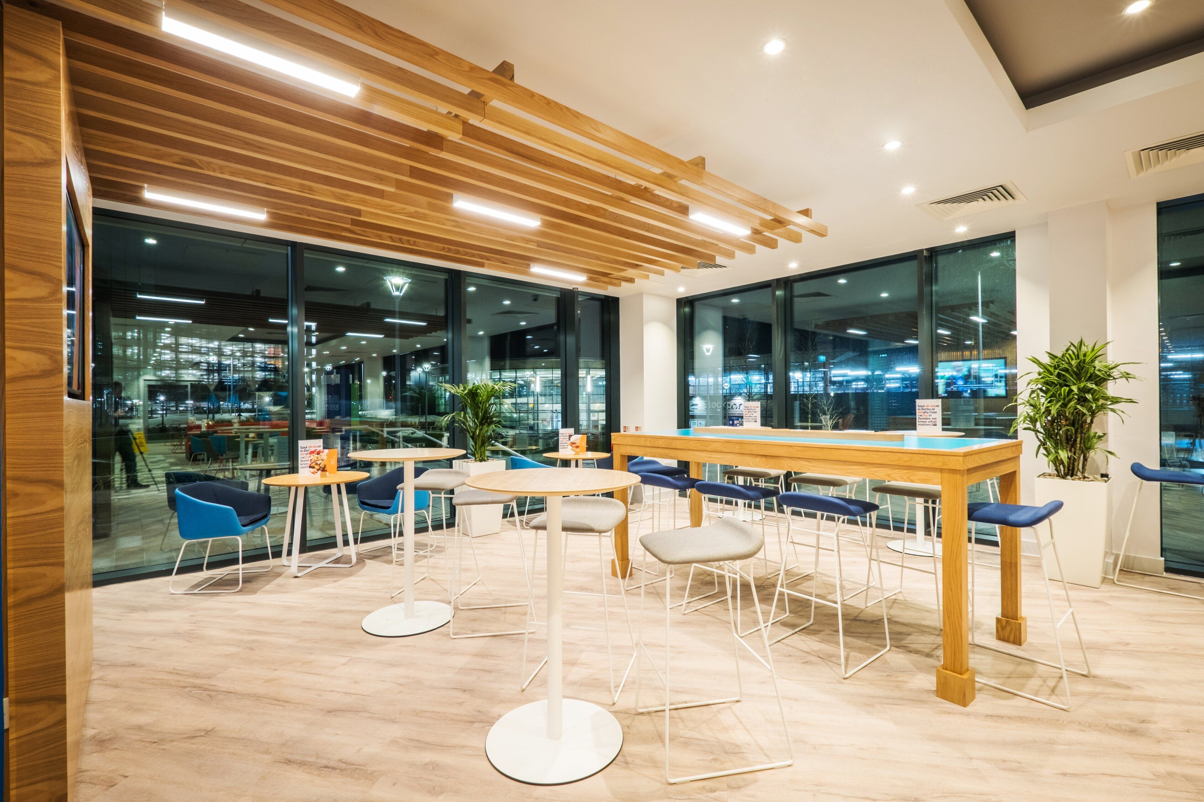 Un espacio de oficina moderno con paneles de techo de madera, paredes de vidrio y una combinación de mesas redondas y cuadradas con sillas.