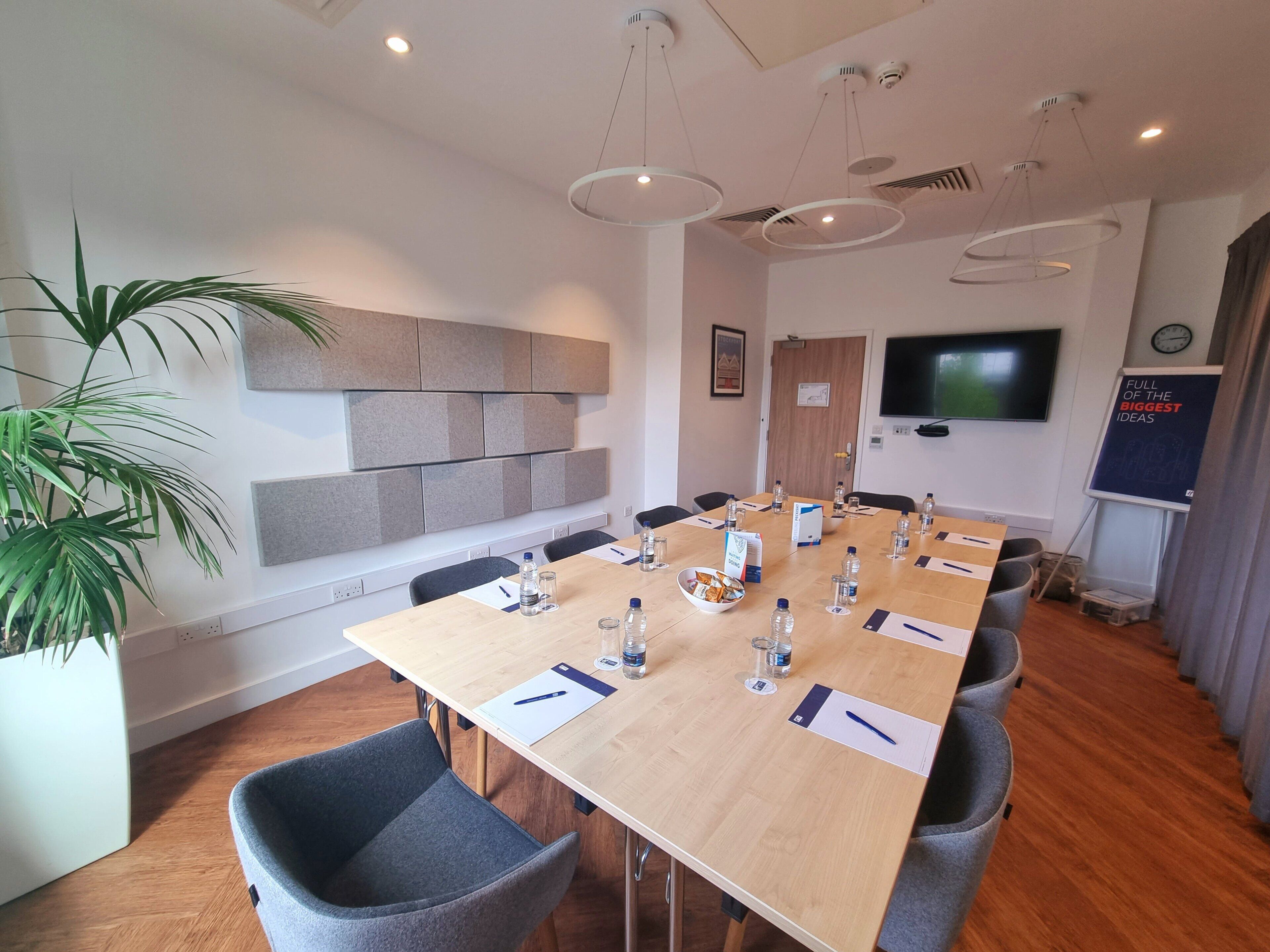 Un salón de conferencias con una larga mesa, sillas, una planta, un televisor y una pizarra.