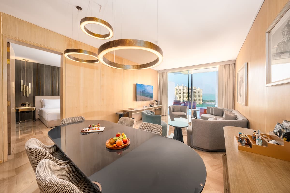 A modern hotel room with a large dining table, comfortable seating, a TV, and a city view.