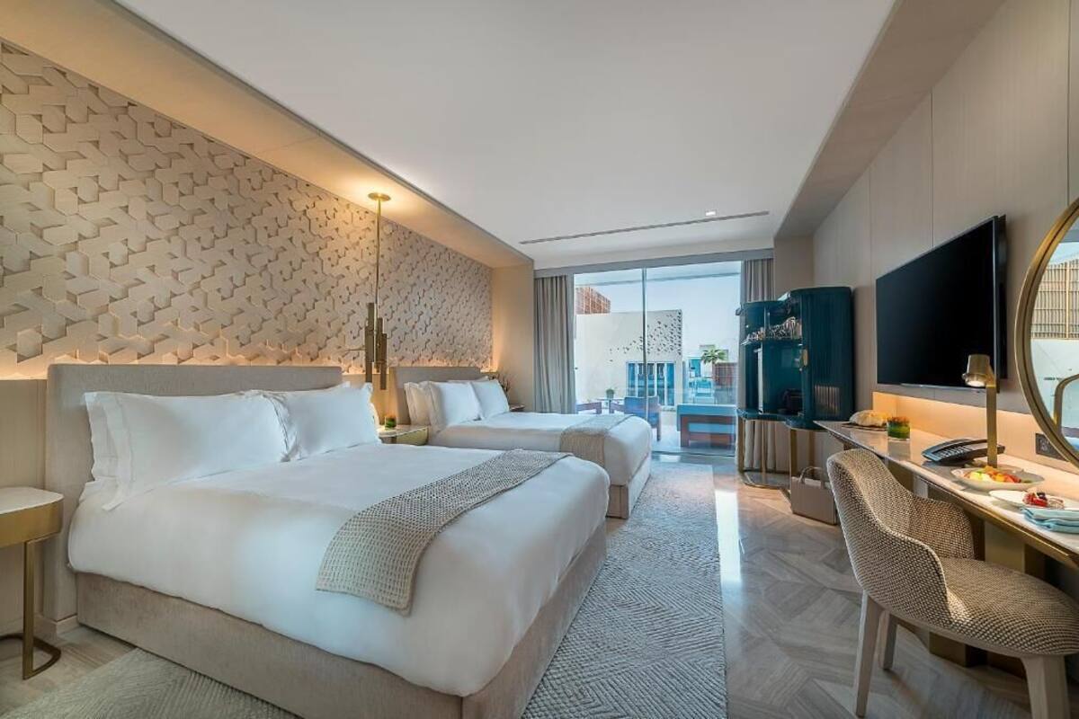 A modern hotel room with two beds, a desk with a chair, a TV, and a large window offering a city view.