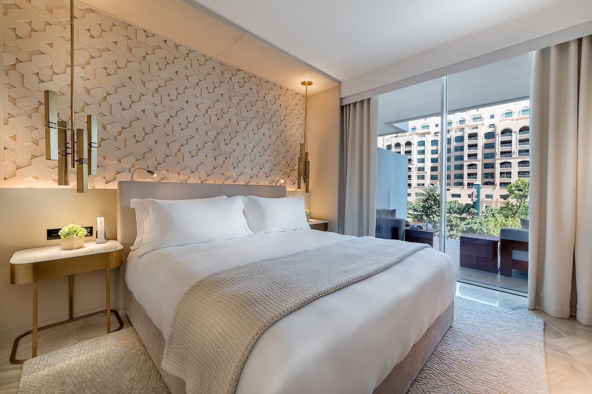 A modern hotel room with a large bed, a bedside table with a lamp, a wall with a geometric pattern, and a view of a cityscape through large windows.
