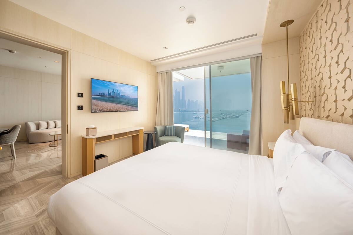 A modern hotel room with a large bed, a desk, a chair, and a view of a cityscape through large windows.