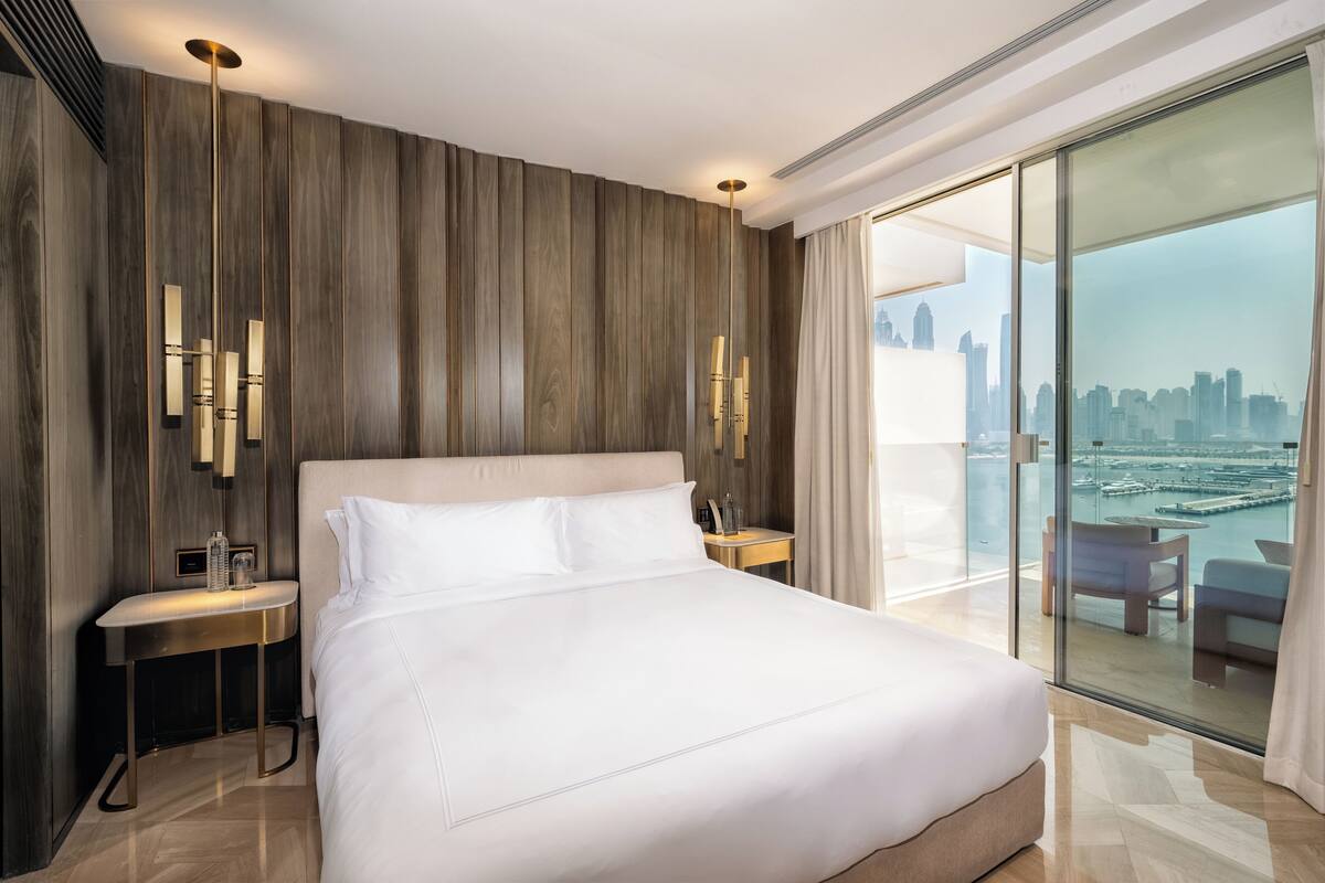 A modern hotel room with a large bed, a nightstand, a lamp, and a view of a cityscape through a glass door.