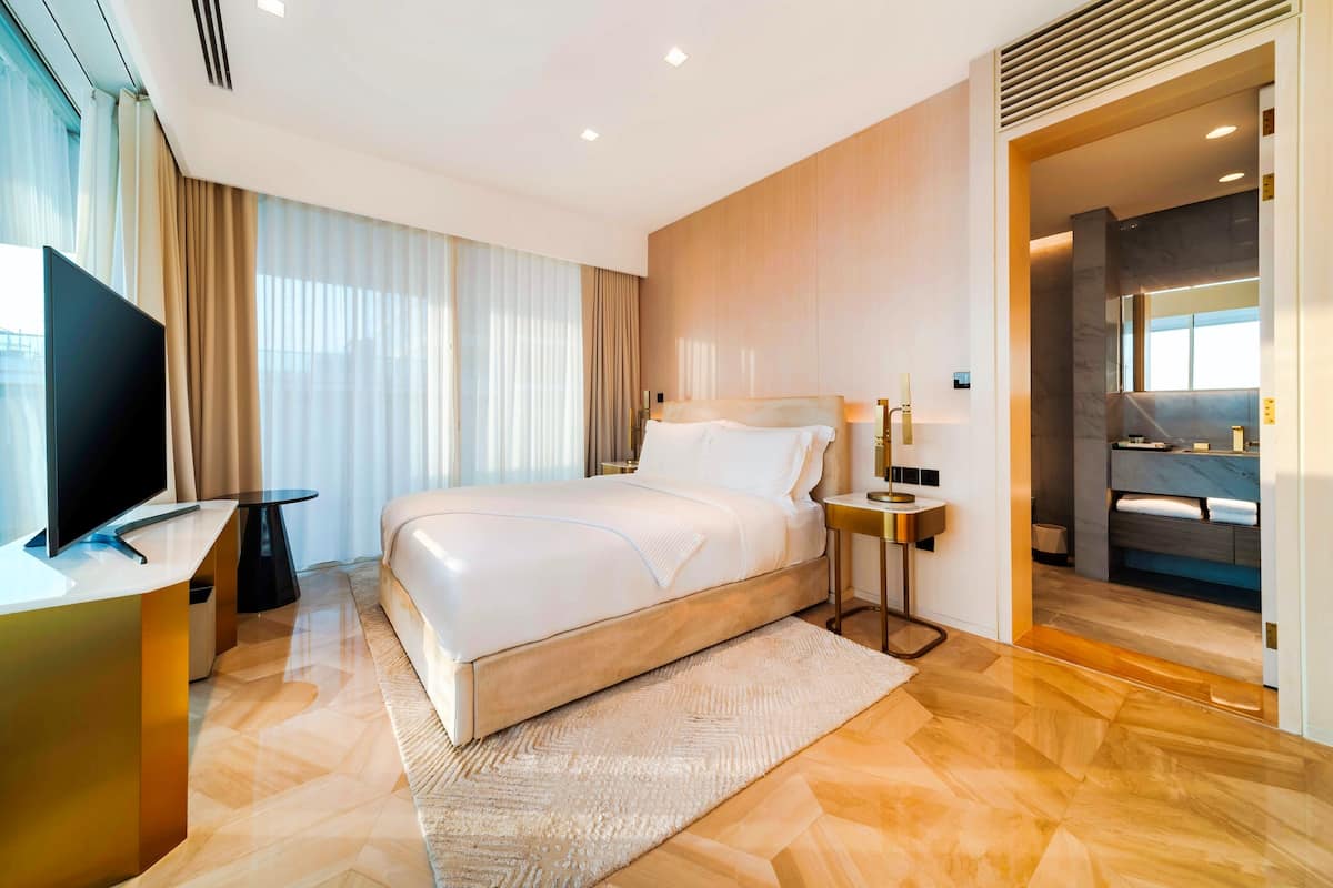 A modern hotel room with a large bed, a TV, a desk, and a bathroom visible through an open door.
