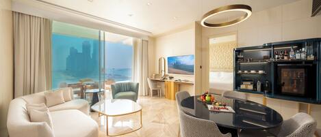 A modern hotel room with a large sofa, a dining table with chairs, a minibar, and a city view.