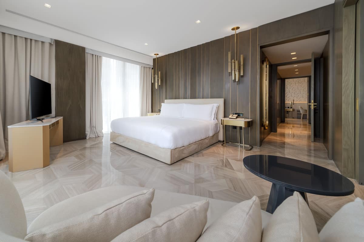 A modern hotel room with a large bed, a TV, a desk, and a dining area.
