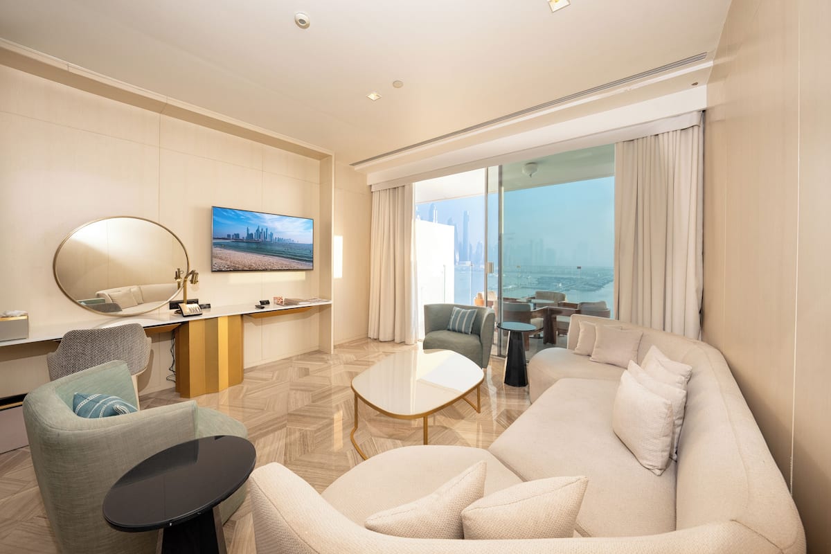 A modern hotel room with a large sofa, a small table, a chair, a desk with a computer, and a large window offering a city view.