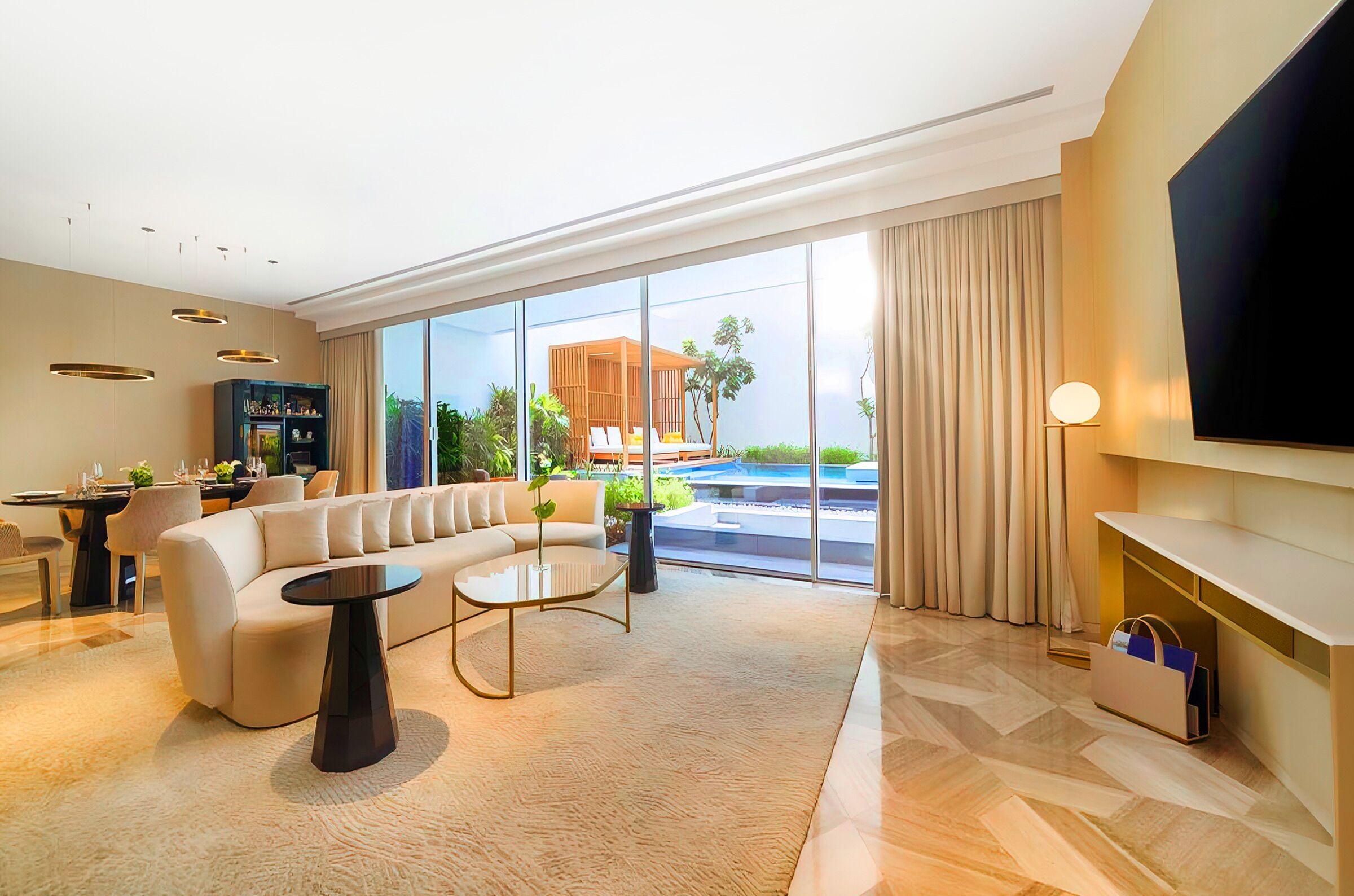 A modern living room with a sofa, coffee table, and TV, large windows with a view of a pool and outdoor seating.