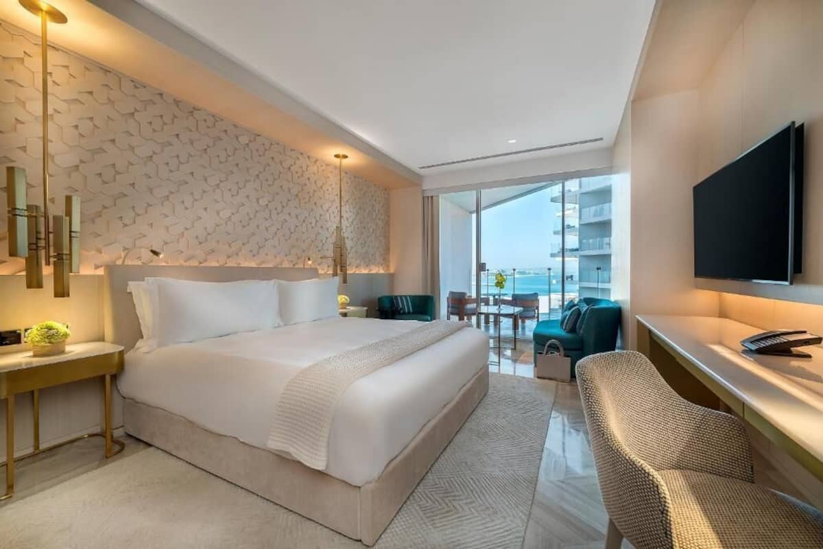 A modern hotel room with a large bed, a desk, a chair, and a TV. There is a balcony with a view of the city.