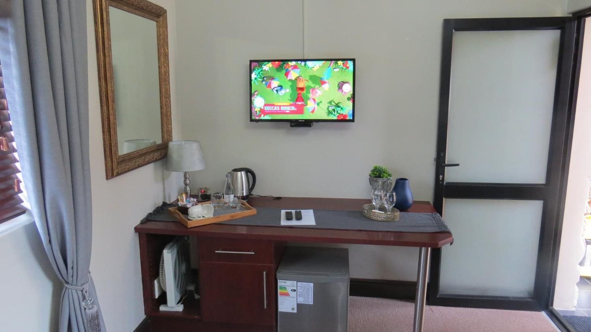 Ein Hotelzimmer mit Schreibtisch, Fernseher, Spiegel, kleinem Kühlschrank und einer Tür.