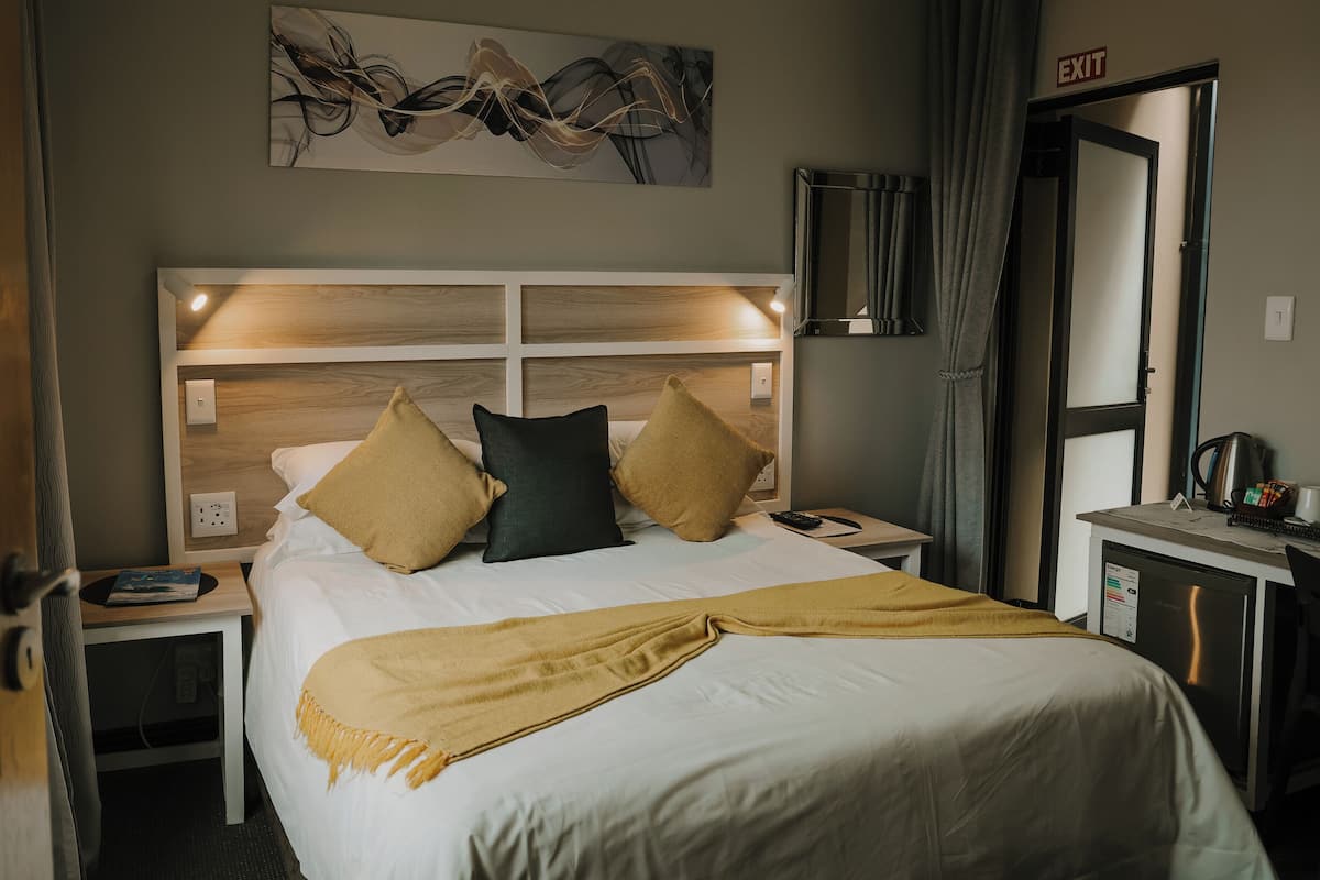 A hotel room with a bed, bedside tables, a nightstand, a wall-mounted artwork, a mirror, and an exit sign.