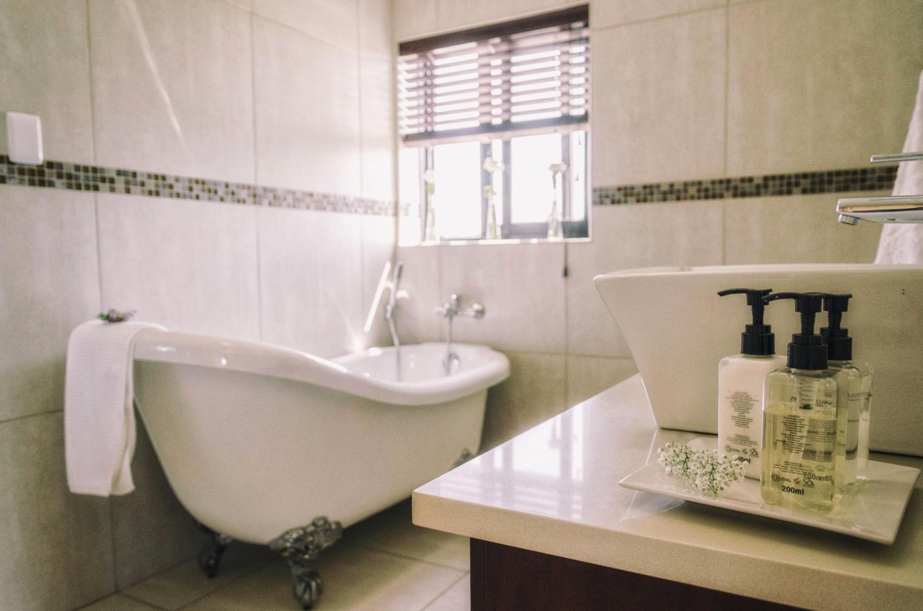 Ein Badezimmer mit Badewanne auf Füßen, Waschbecken und Fenster mit Jalousien. Auf dem Waschbecken stehen zwei Flaschen Flüssigseife.
