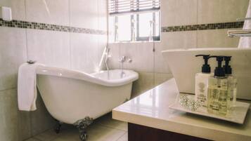 Ein Badezimmer mit Badewanne auf Füßen, Waschbecken und Fenster mit Jalousien. Auf dem Waschbecken stehen zwei Flaschen Flüssigseife.