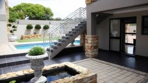 A modern outdoor pool area with a stone fountain, a staircase leading to a covered area, and a view of the surrounding landscape. - Stone Olive Guest House (Jeffreys Bay)