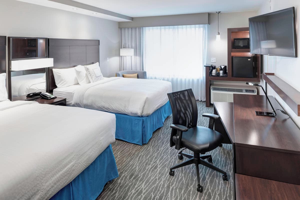 A hotel room with two beds, a desk with a chair, a TV, a microwave, and a window with curtains.