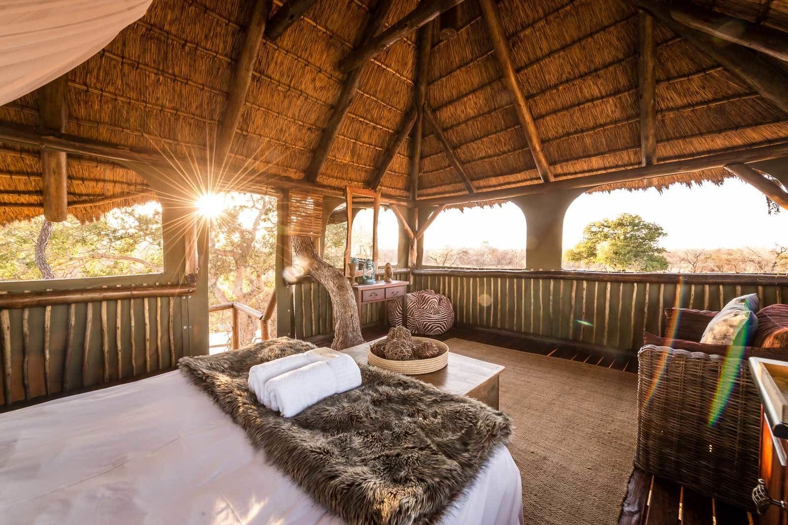 A thatched-roof room with a bed, a small table, and a view of trees.