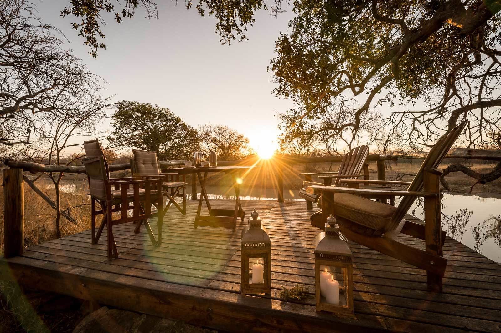 A wooden deck with chairs and tables, overlooking a body of water with the sun setting in the background.