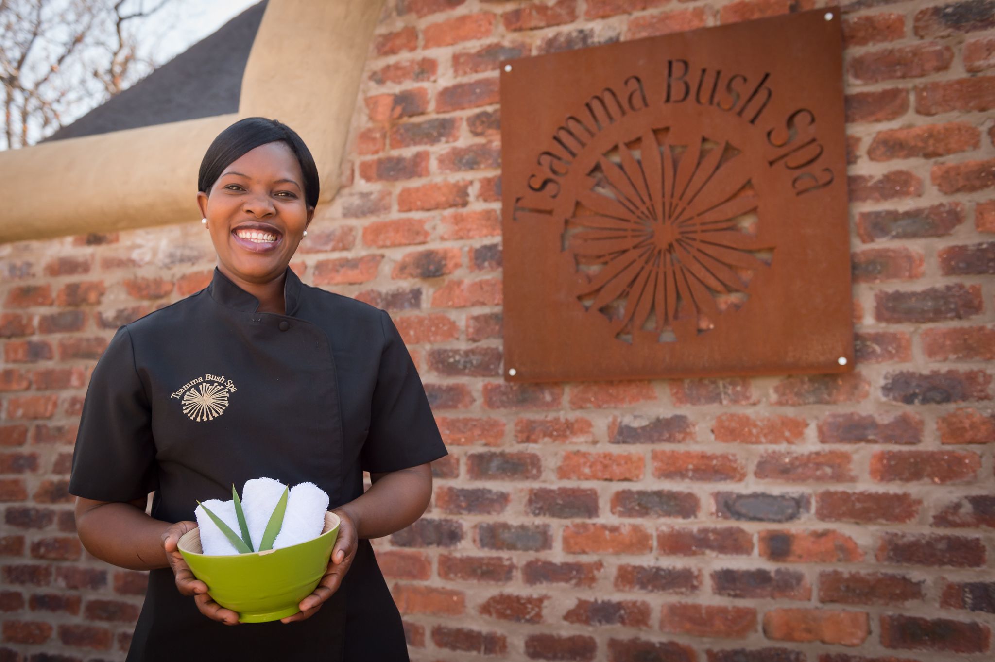 A person holding a green bowl with white towels, standing in front of a brick wall with a sign that reads "Tsamma Bush Spa".