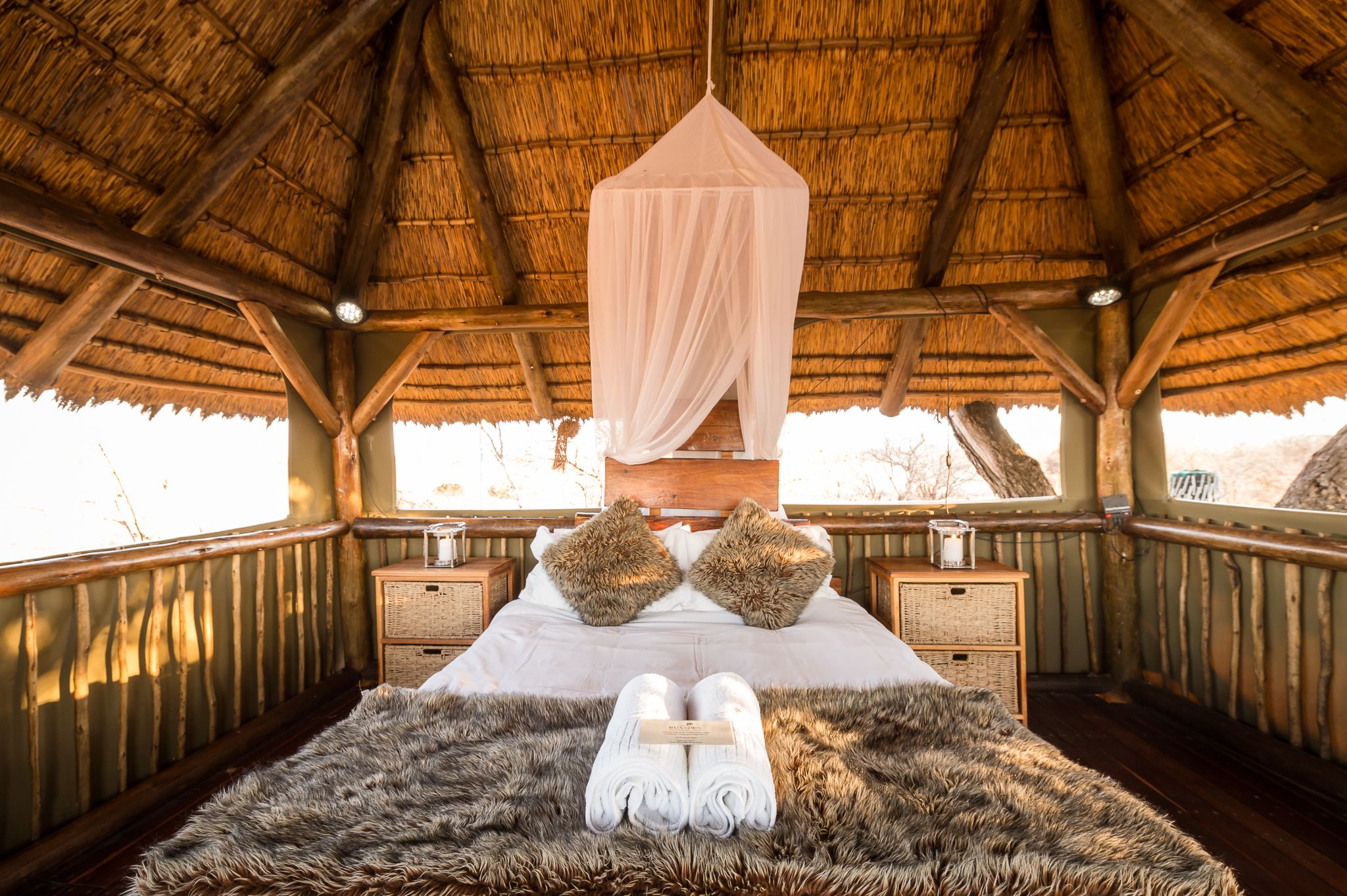 A room with a thatched roof, wooden beams, a bed with white linens, two fluffy pillows, and a fur rug.
