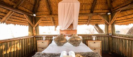 A room with a thatched roof, wooden beams, a bed with white linens, two fluffy pillows, and a fur rug.