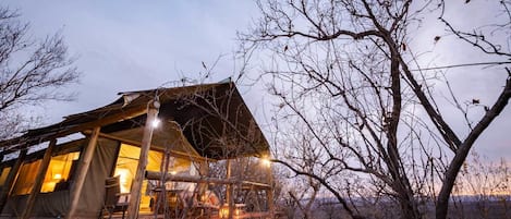 A rustic, tent-like structure with a wooden deck, surrounded by bare trees and rocks, likely a remote accommodation.