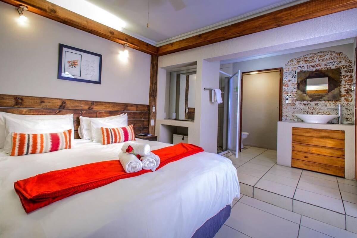 A hotel room with a wooden headboard, a large bed with white bedding, a red throw, and a bathroom with a wooden vanity and a sink.