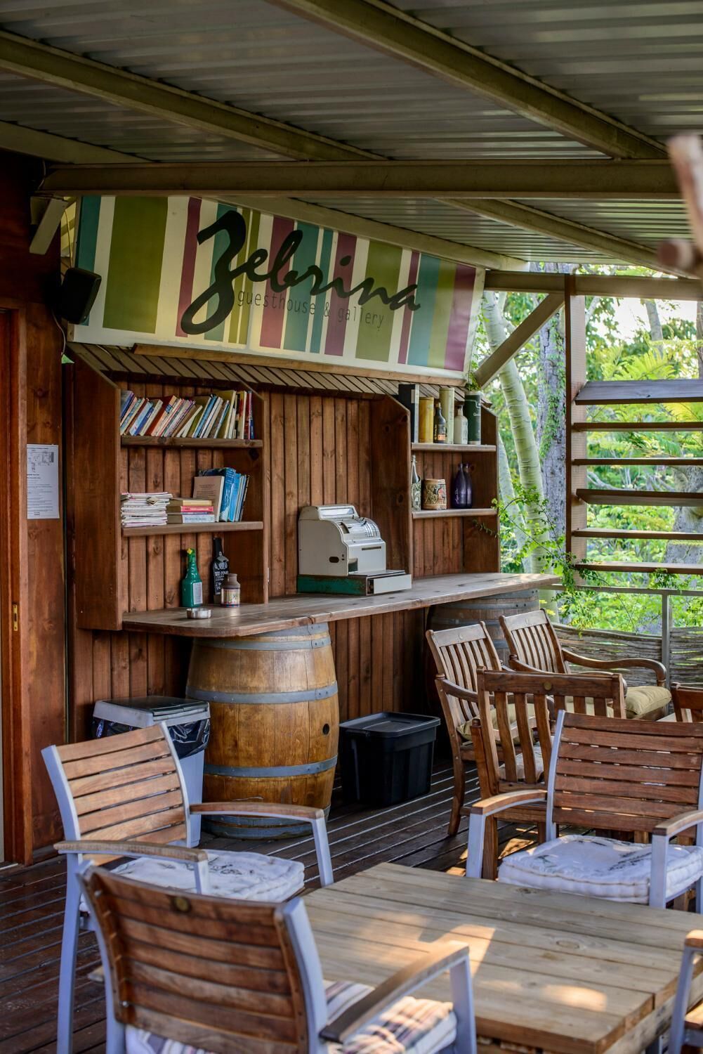 Uma área de estar rústica ao ar livre, com mobiliário de madeira, um bar com uma placa listrada e uma estante cheia de livros.