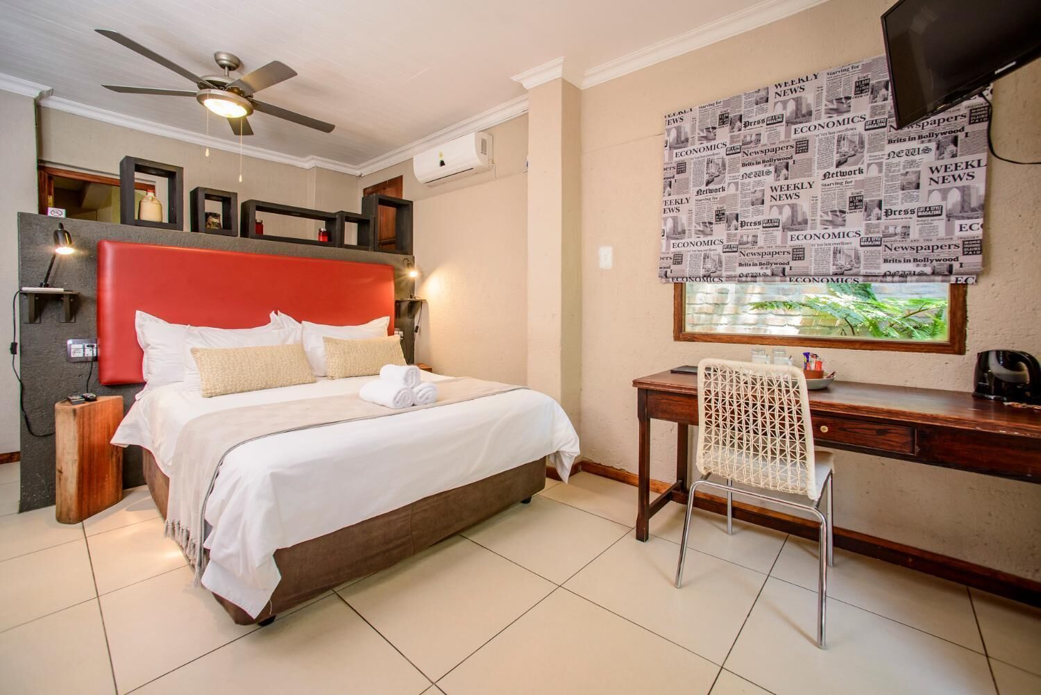 A hotel room with a bed, a desk, a chair, a ceiling fan, and a wall with newspaper clippings.