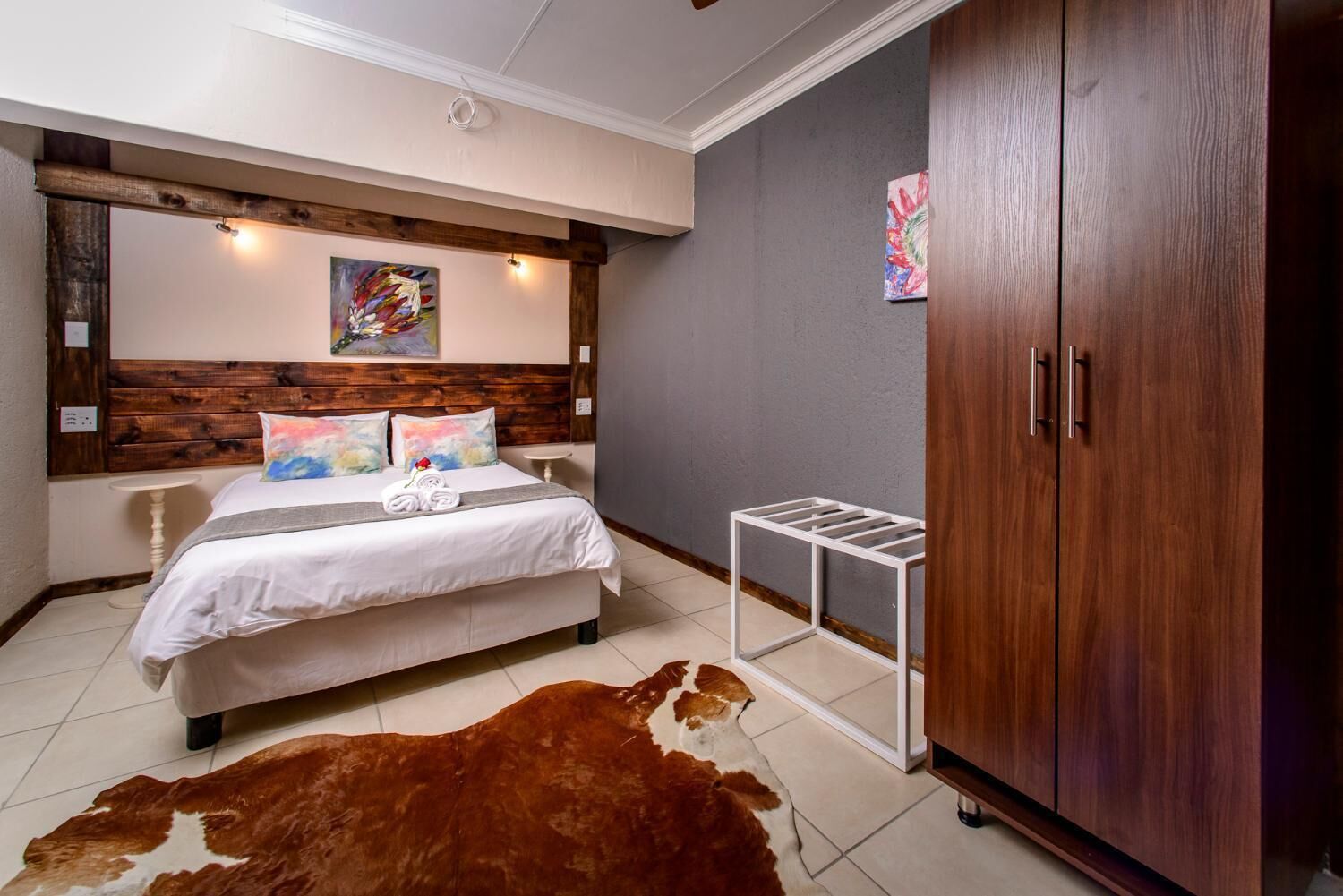 A bedroom with a wooden headboard, a bed with white linens, a cowhide rug, a white metal side table, and a wooden wardrobe.