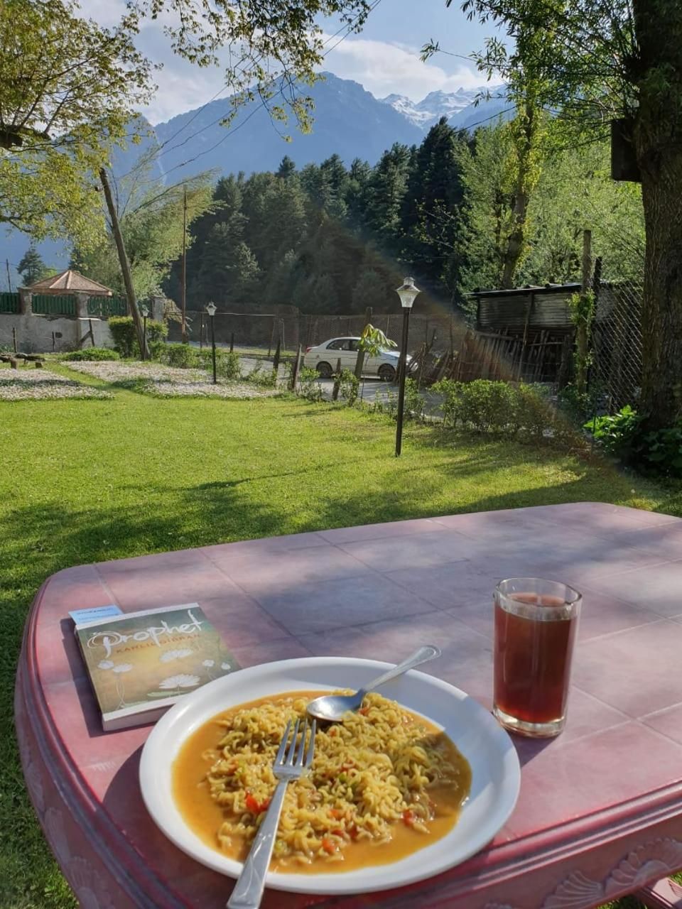 Une table dressée avec un repas, un livre et une boisson, le tout dans un cadre extérieur pittoresque.