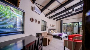 Una habitación con una mesa de comedor, sillas, un sofá, una cama y una silla roja. Hay ventanas con persianas, un ventilador de techo y un aire acondicionado instalado en la pared.