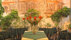 A dining area with a table set for two, surrounded by chairs. There is a vase with red flowers on the table. - Mirador del Monasterio (Arequipa)