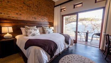 A bedroom with a large bed, a brick wall, a view through a glass door, and a small table with chairs outside.
