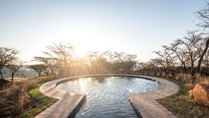 A circular pool with a brick border, surrounded by grass and trees, with the sun setting in the background. - The Hilton Bush Lodge (Hilton)