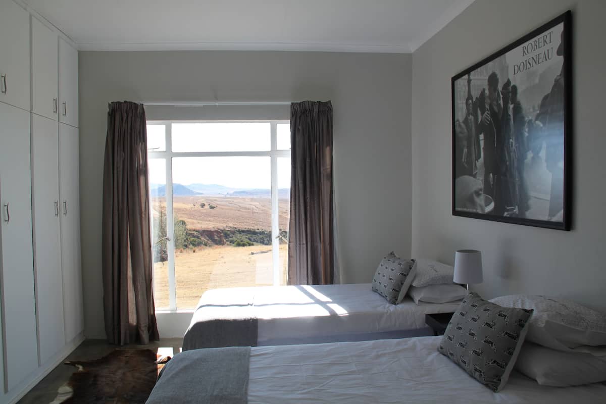 A bedroom with two beds, a large window with curtains, a mounted picture, and a wardrobe.