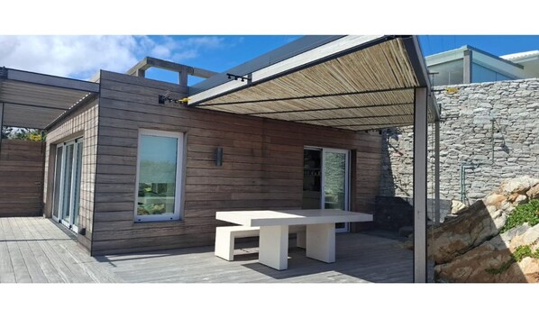 Un patio moderno al aire libre con piso de madera, pared de piedra y mesa blanca.