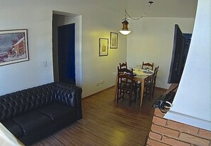 A living room with a black leather sofa, a dining area with wooden chairs and a table, and a TV mounted on the wall.