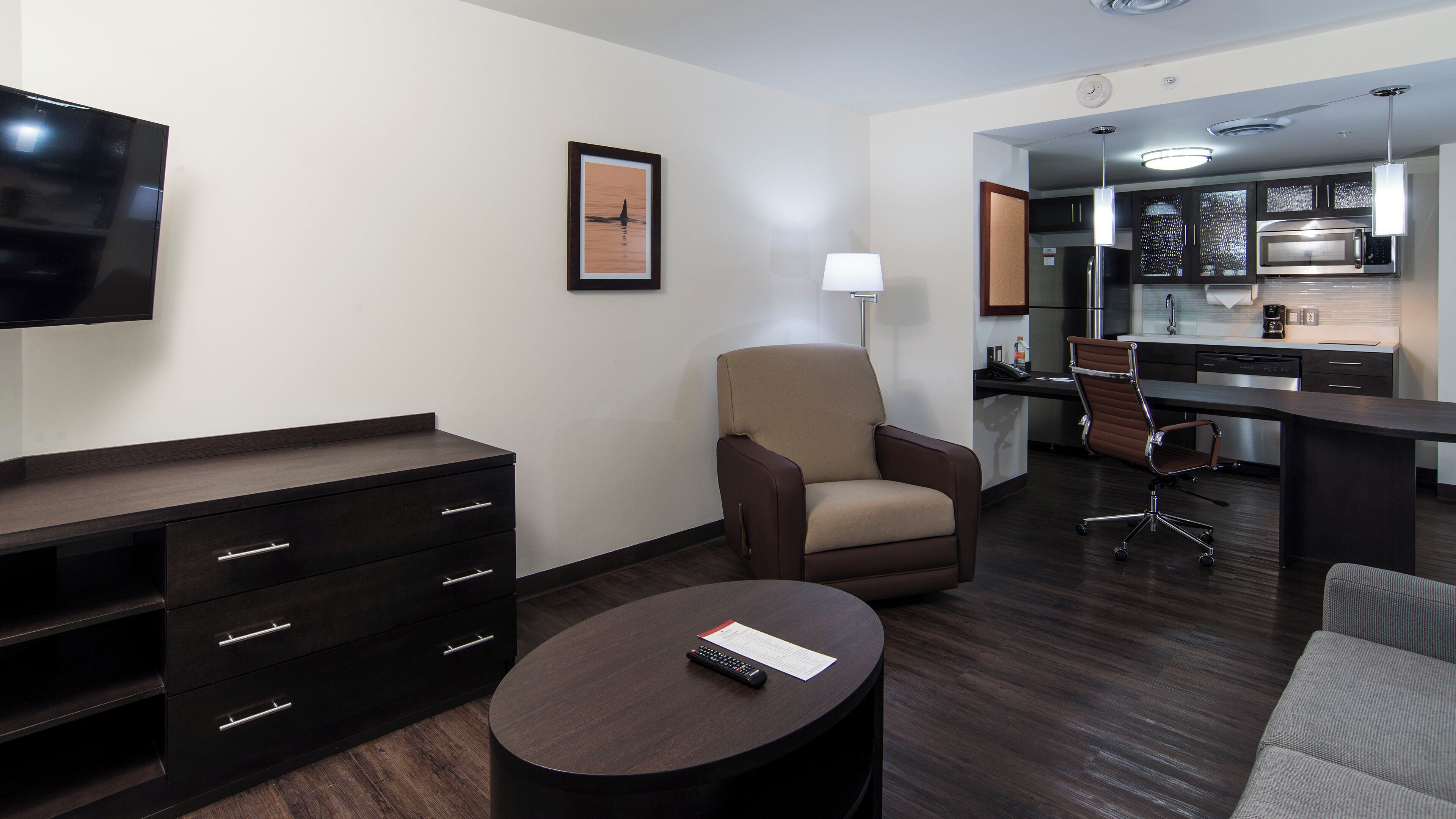 A modern hotel room with a flat-screen TV, a sofa, a chair, a small round table with a remote control, and a kitchen area with a microwave and coffee maker.