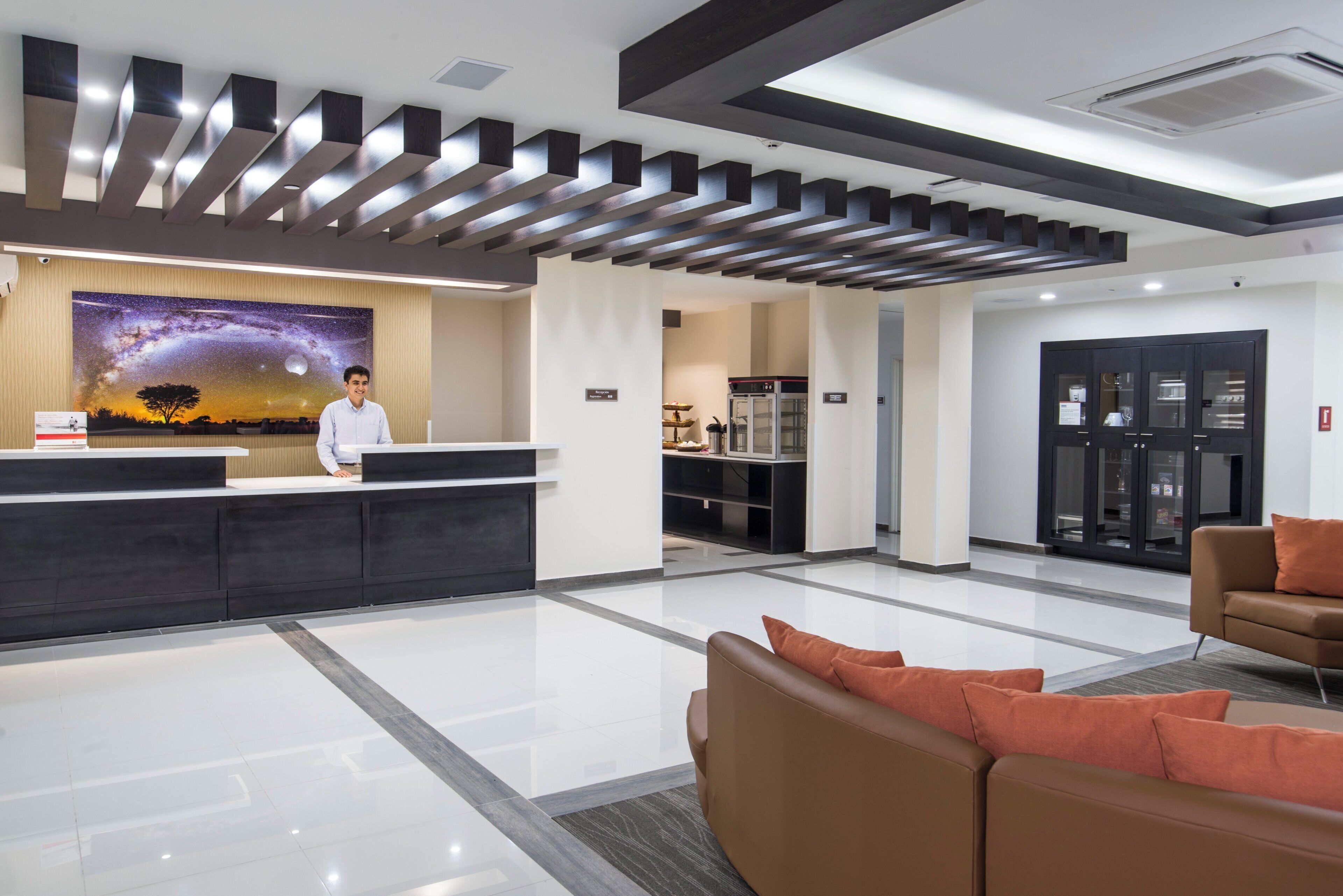 A modern reception area with a receptionist, a large wall mural, a kitchen area, and seating.