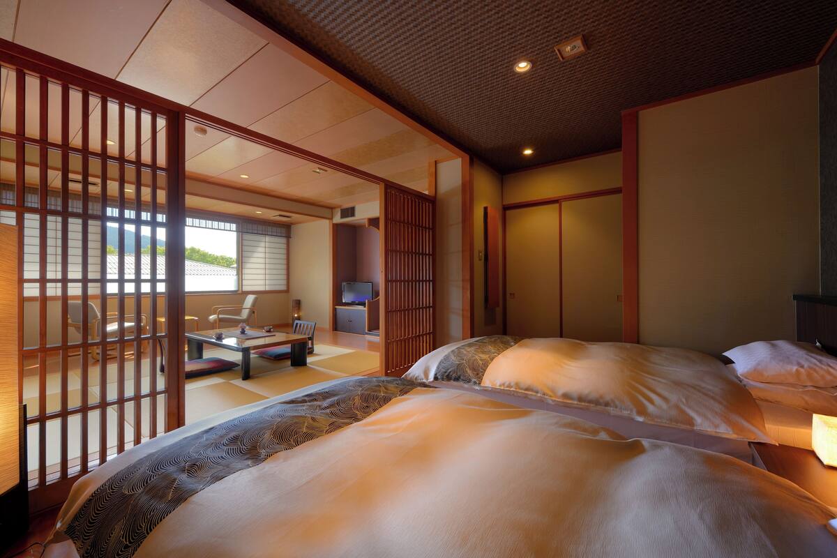 Ein traditioneller japanischer Raum mit Tatami-Bodenbelag, einem niedrigen hölzernen Bett mit gemusterten Textilien und einer Schiebetür zu einem Sitzbereich mit Tisch und Stühlen.